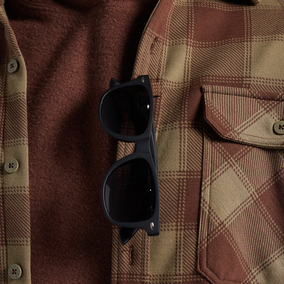 Close-up of sunglasses hanging on the sunglass loop of a brown and tan plaid flannel shirt over a rust-brown sweater.