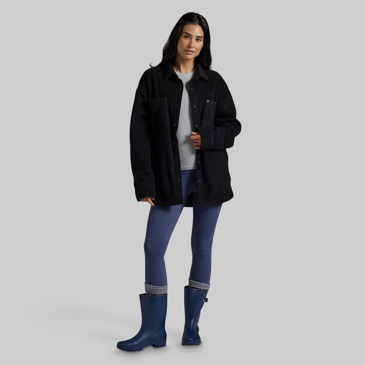 A woman modeling a black sherpa fleece jacket over a gray top, blue leggings, and blue rain boots against a solid gray studio background.
