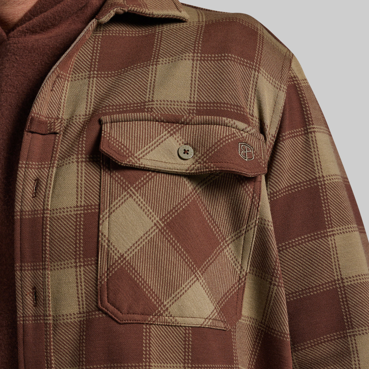 Close-up of a brown and beige plaid flannel shirt jacket with a chest pocket and snap tab, layered over a brown hoodie against a light gray background.