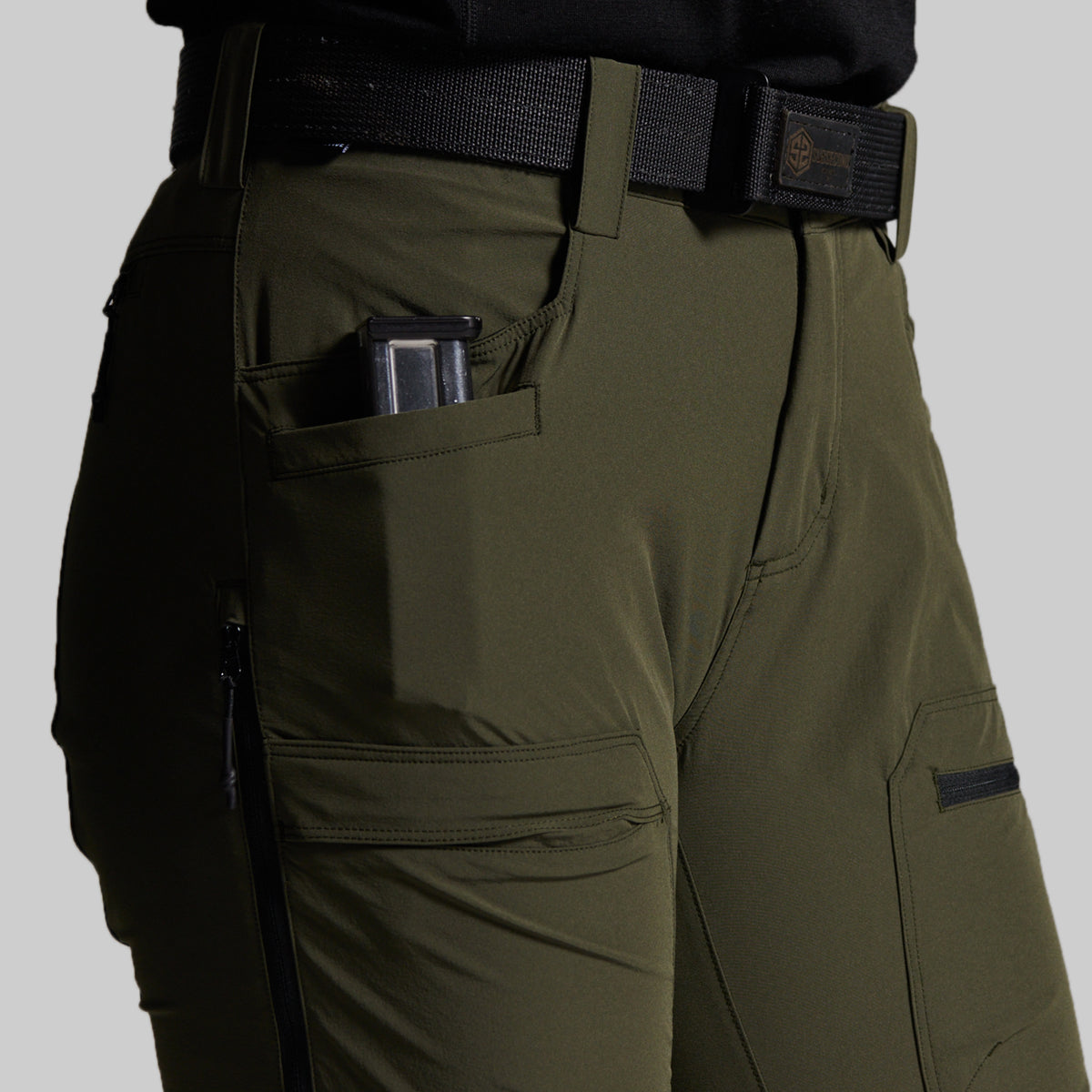 Close-up of olive green outdoor pants worn with a black belt, showing side pockets, a zippered pocket, and a handgun magazine in the side pocket against a light gray background.