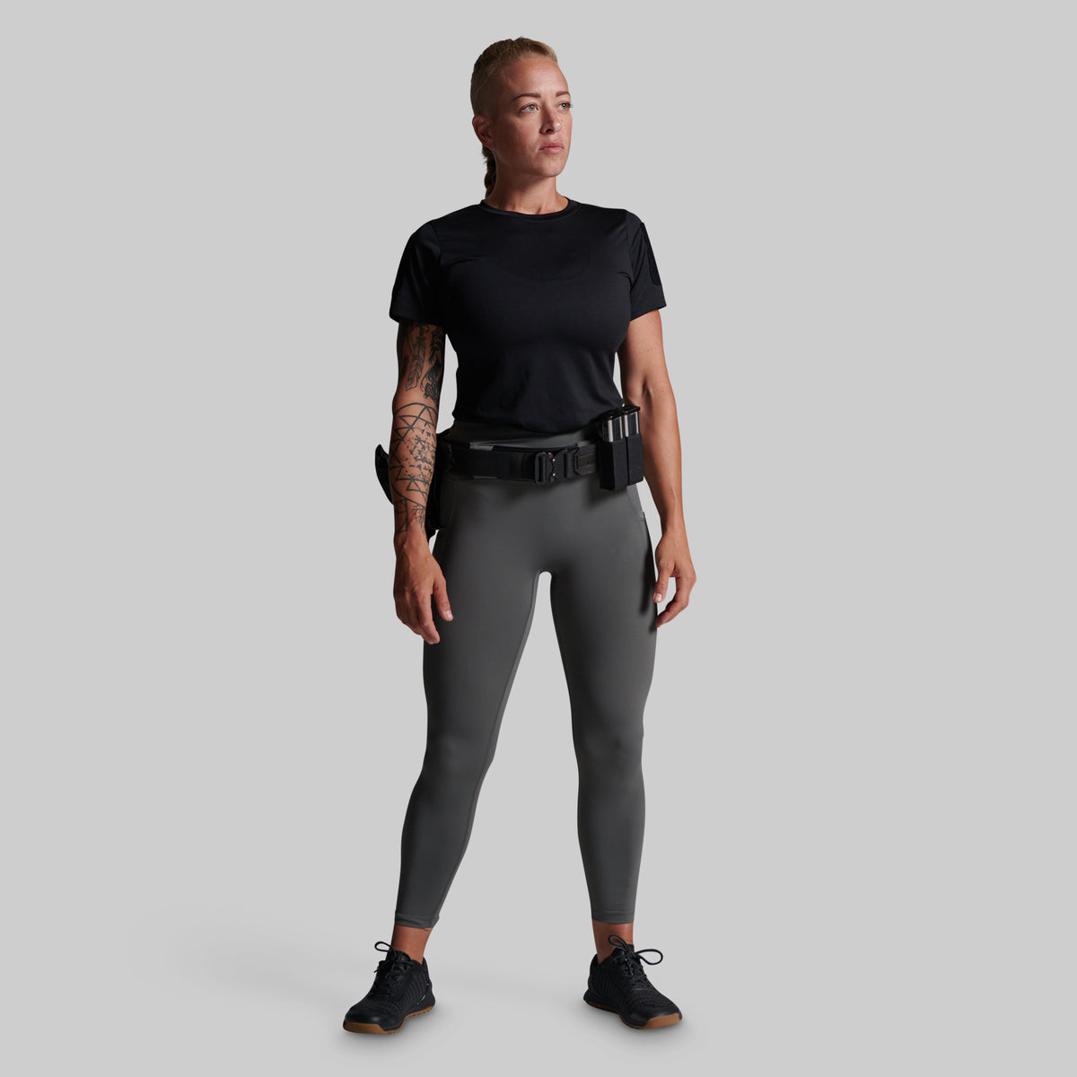 A woman modeling a black short-sleeve tactical shirt and dark gray leggings with a black utility belt and holster, standing against a solid gray background.