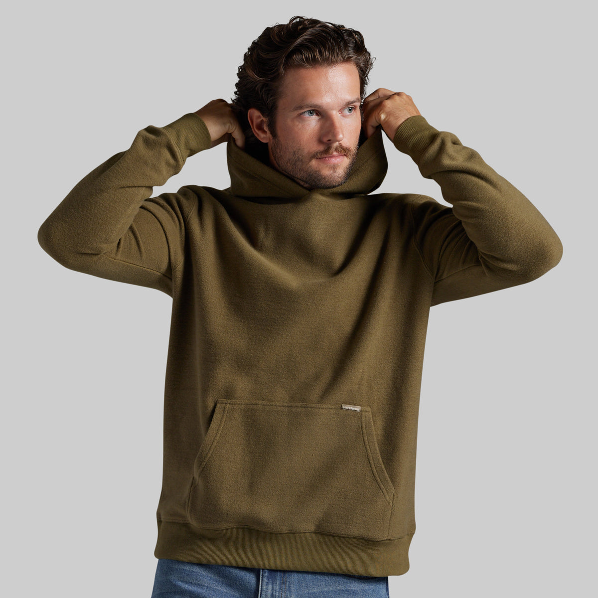 A man wearing an olive green fleece pullover hoodie with a front kangaroo pocket, shown from the waist up against a solid gray background.