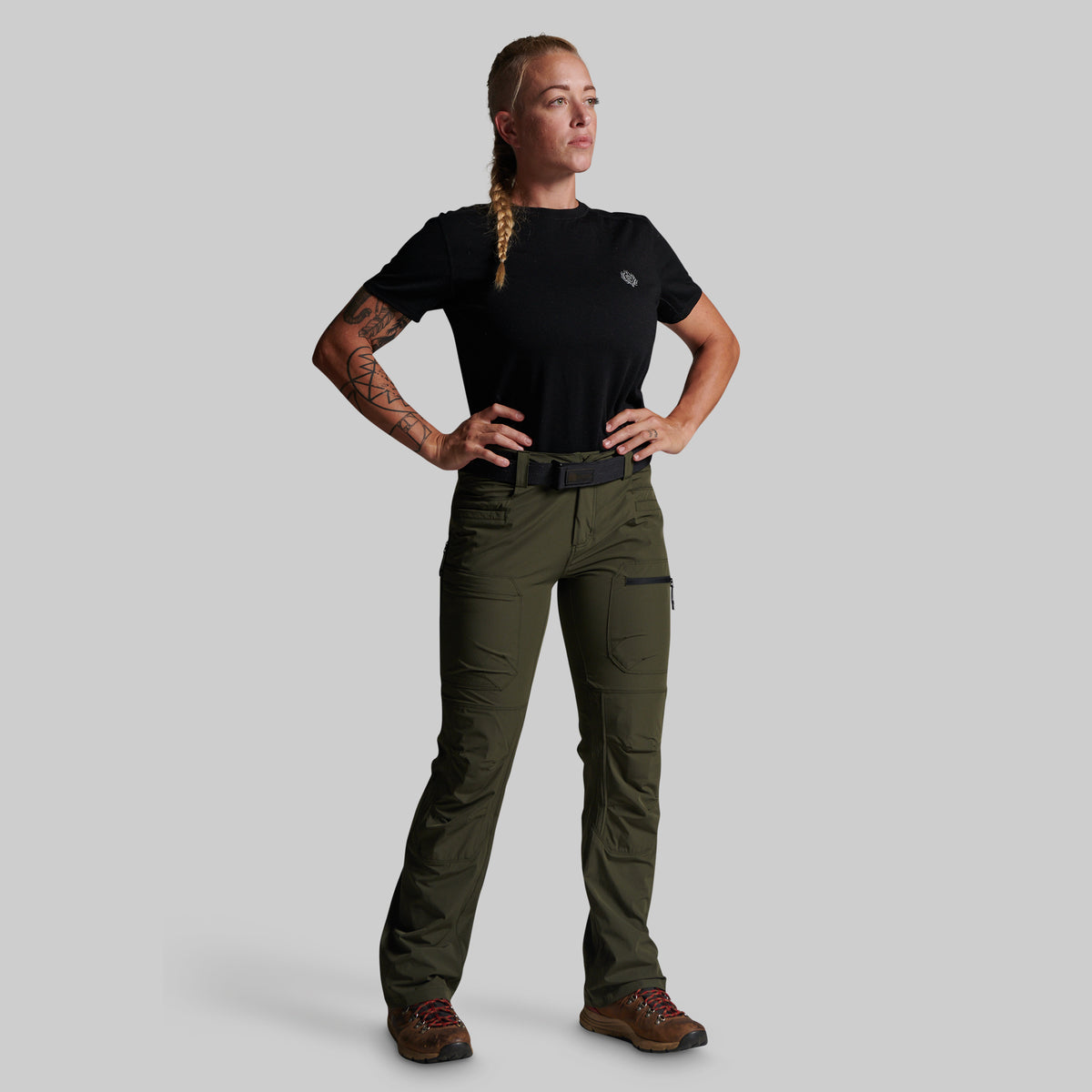 A woman modeling a pair of olive green outdoor cargo pants with a black t-shirt and brown hiking boots, standing against a solid gray background.