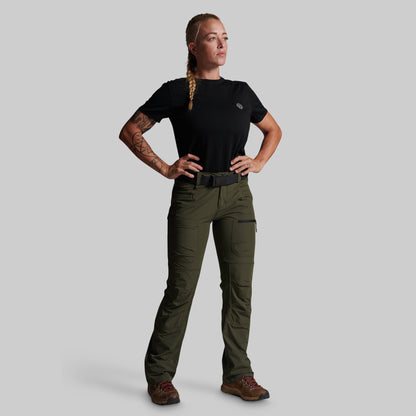 A woman modeling a pair of olive green outdoor cargo pants with a black t-shirt and brown hiking boots, standing against a solid gray background.