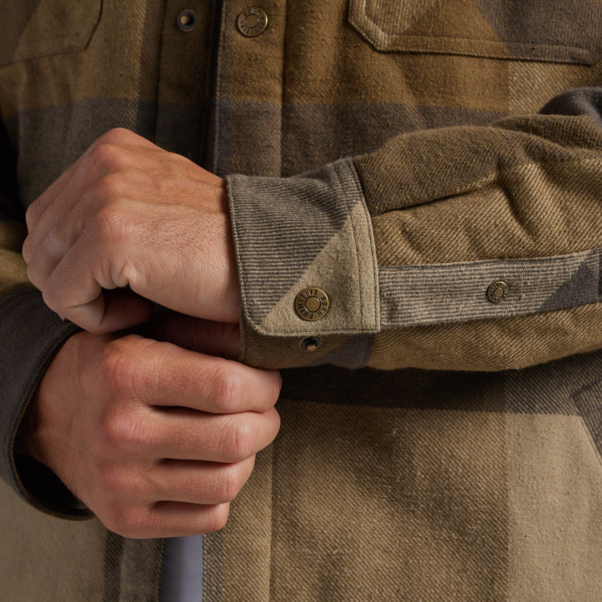 Close-up of a person's hands adjusting the cuff of a brown and tan plaid flannel jacket, showing the fabric and snap-button cuff.