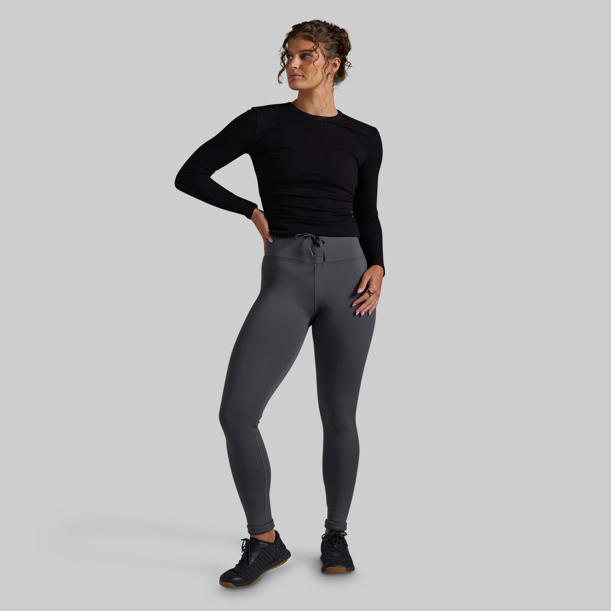 A woman modeling dark gray athletic leggings and a black long-sleeve top against a solid gray studio background.