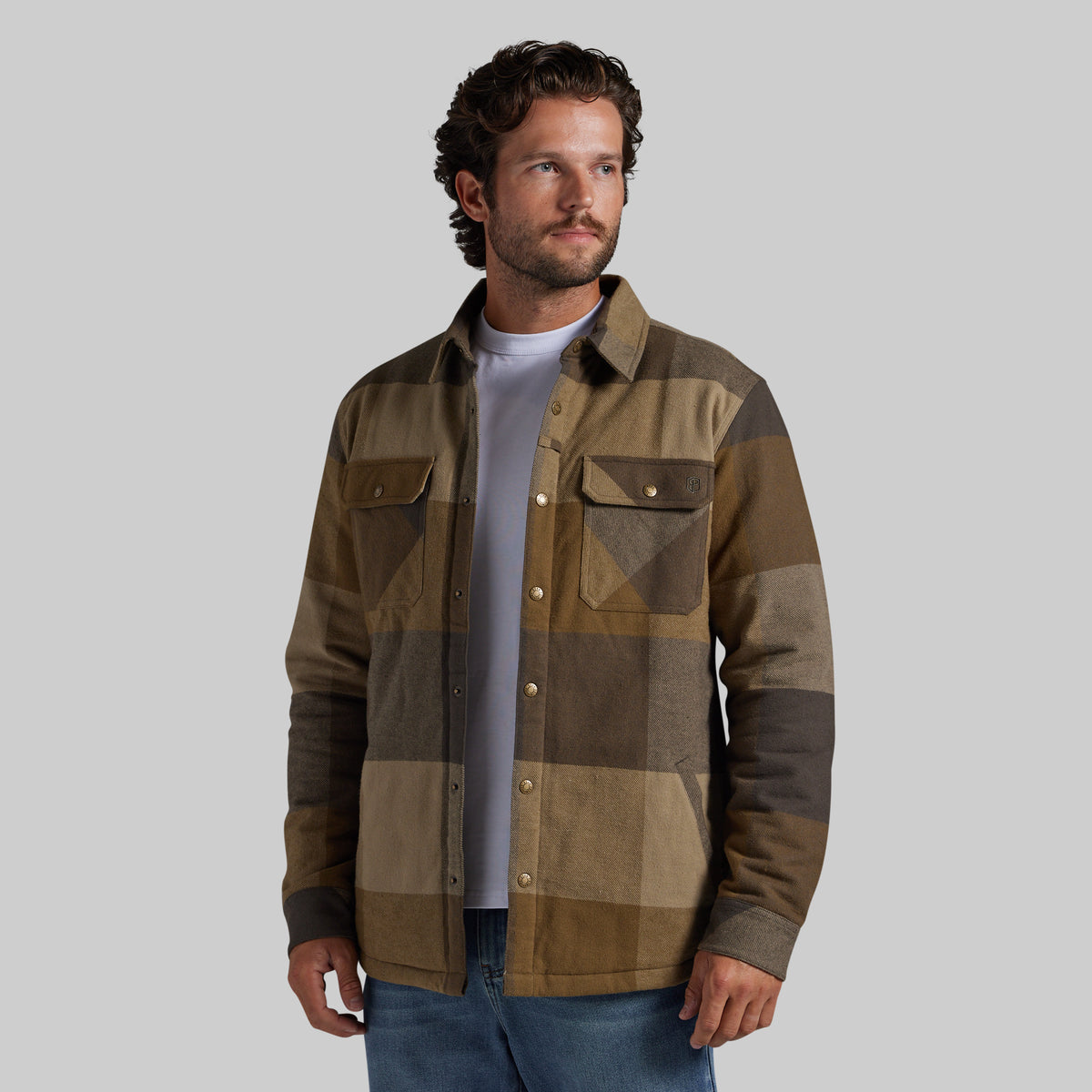 A man wearing a brown and tan patchwork button-up jacket over a light gray t-shirt and blue jeans, photographed against a solid gray studio background.