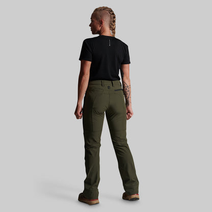 A woman shown from the back wearing olive green outdoor pants, a black t-shirt, and brown boots against a solid gray studio background.