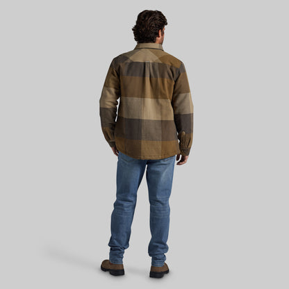 A man viewed from behind wearing a brown and beige checkered flannel jacket, blue jeans, and brown boots against a solid gray background.