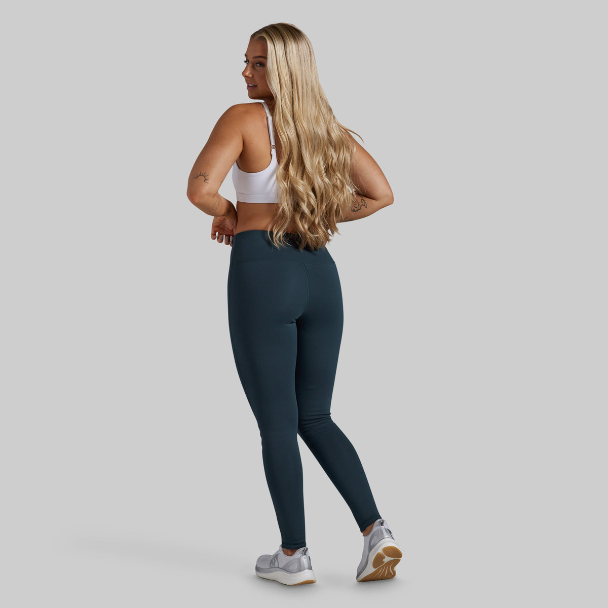 A woman is shown from the back wearing dark teal high-waisted leggings, a white sports bra, and gray running shoes against a solid light gray background.