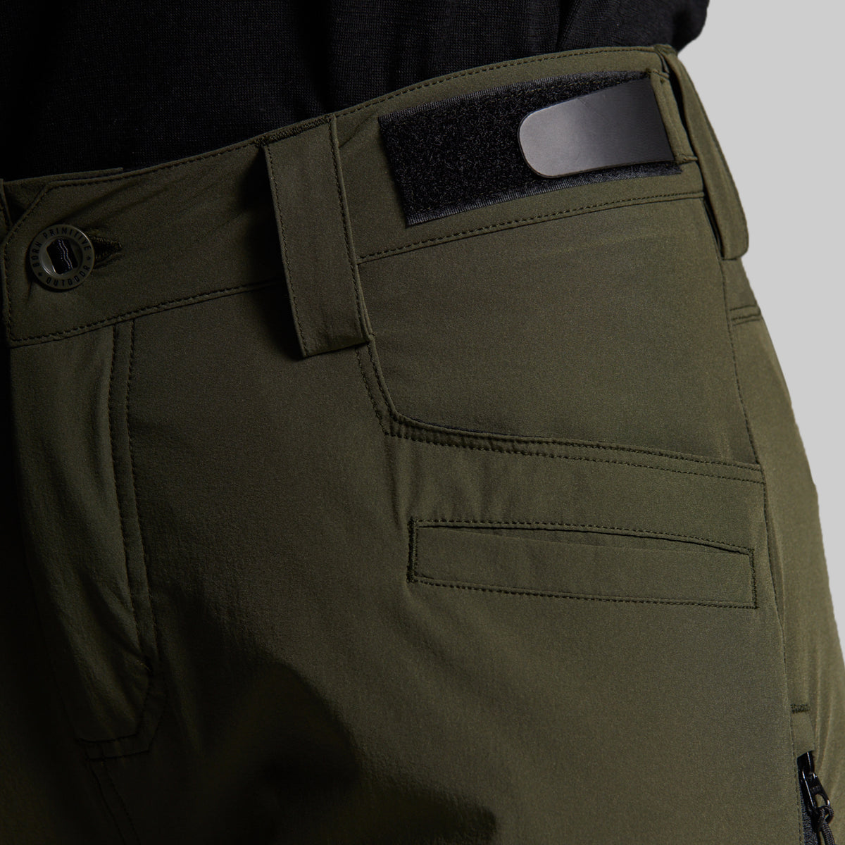 Close-up of olive green outdoor pants showing the waistband, belt loop with a black insert, button closure, and front pocket stitching.