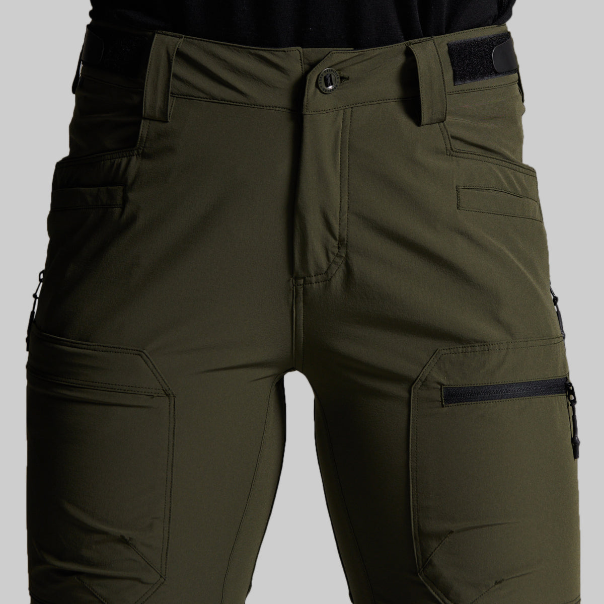 Close-up of olive green outdoor cargo pants worn by a model, showing the front waistband, button closure, belt loops, and side cargo pockets with black zippers against a neutral gray background.
