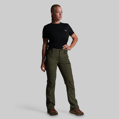 A woman modeling olive green cargo pants with a black t-shirt and brown hiking boots, standing against a solid gray background.