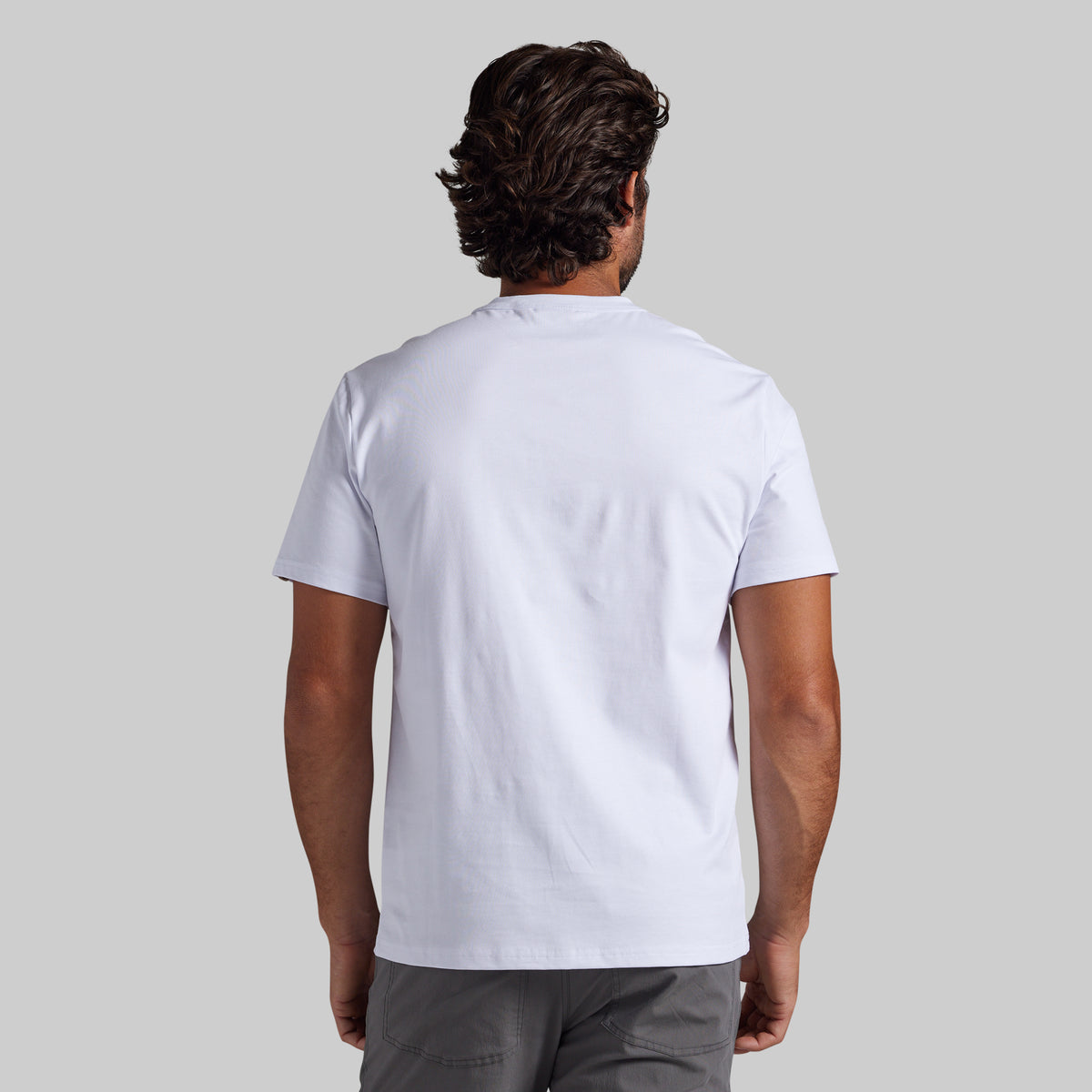 A man viewed from behind wearing a plain white short-sleeve t-shirt and gray pants, set against a solid light gray background.