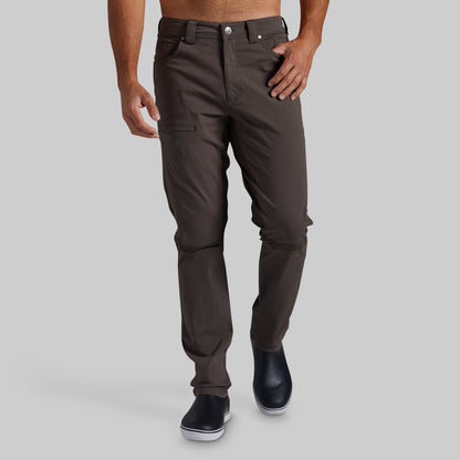 A man wearing dark brown slim-fit pants with utility pockets, shown from the waist down against a solid gray background.