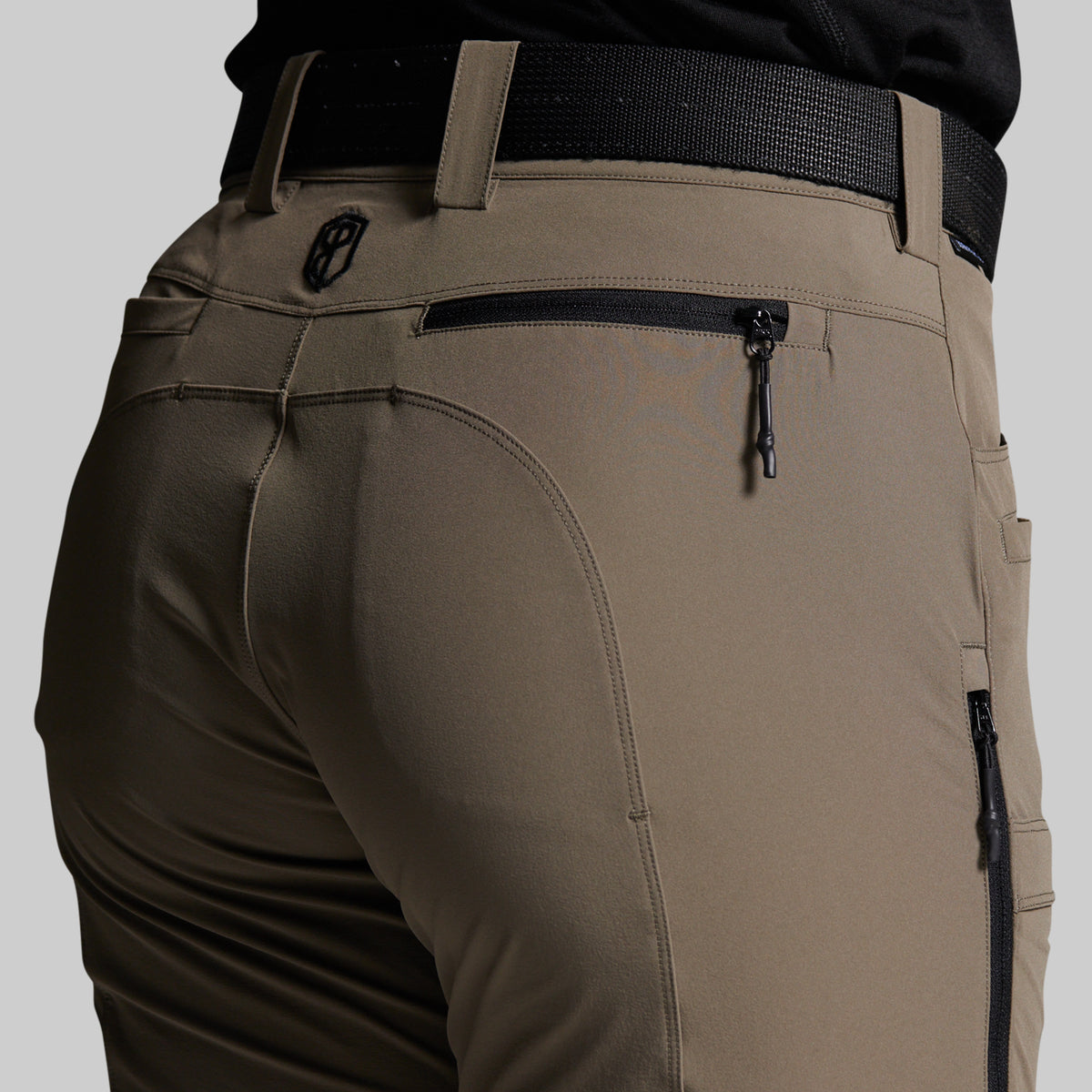 Close-up of the back waist and pockets of storm grey outdoor pants worn with a black belt, showing zippered pockets and a small circular logo emblem.