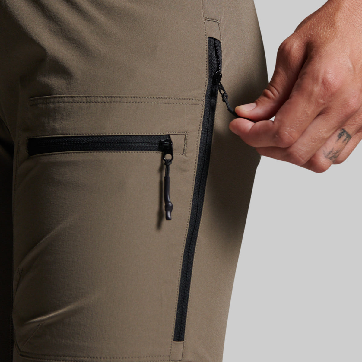 Close-up of storm grey outdoor pants showing a side zippered pocket and a hand pulling the zipper tab.