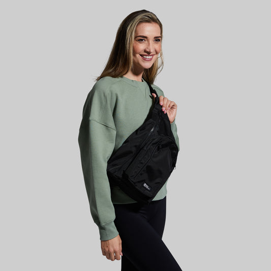 Woman wearing a green sweatshirt and black sling bag against a gray background
