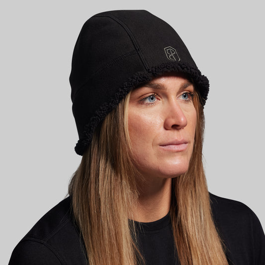 Person wearing a black beanie with a logo on a gray background