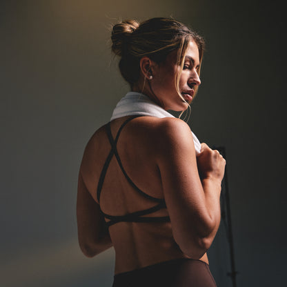 A woman in a brown strappy sports bra and dark leggings, with a white towel around her neck, shown from the back in a dimly lit studio setting.