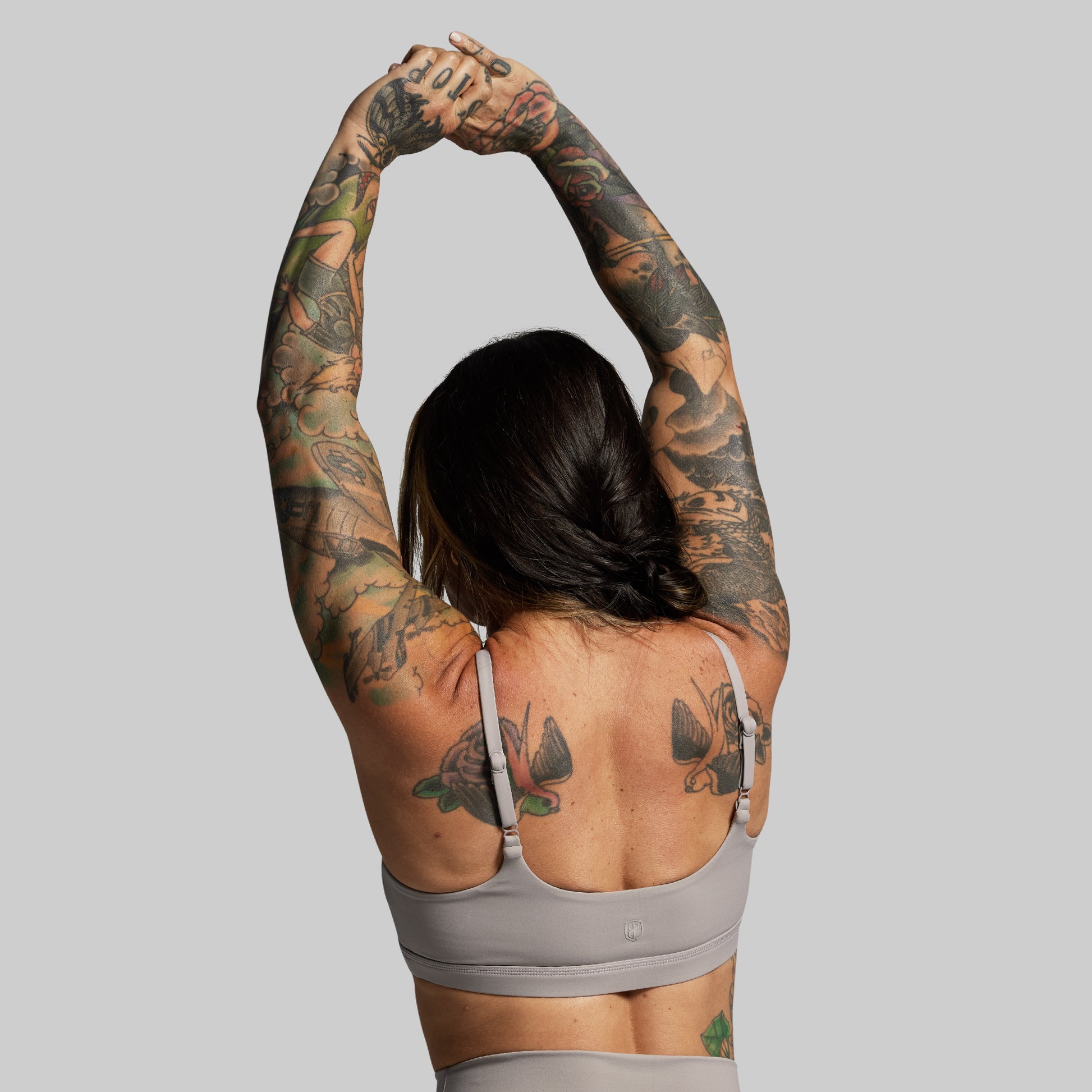 Person with tattoos stretching arms wearing gray sports bra against a gray background