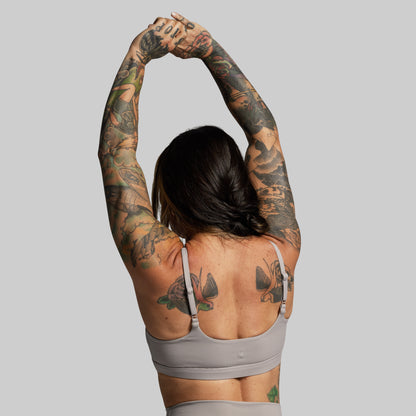 Person with tattoos stretching arms wearing gray sports bra against a gray background