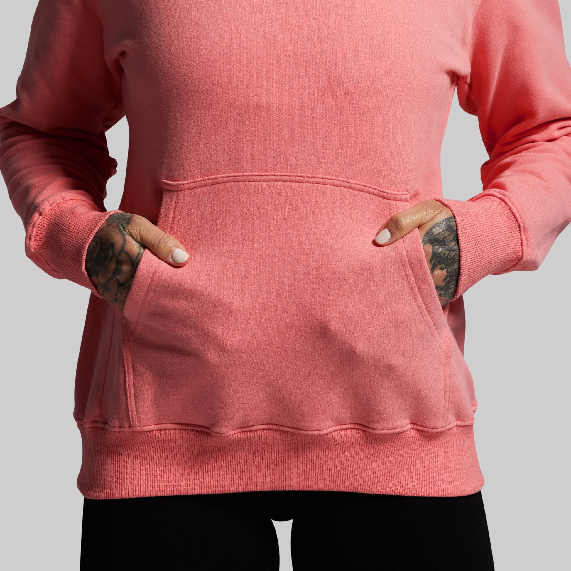 Person wearing a pink hoodie with tattoos on arms against a gray background