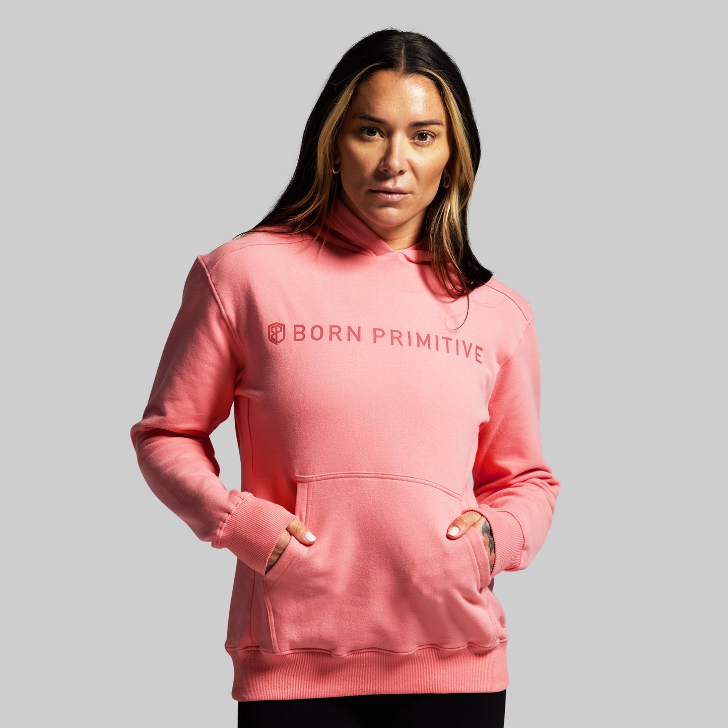 Person wearing a pink hoodie with 'Born Primitive' logo on a gray background
