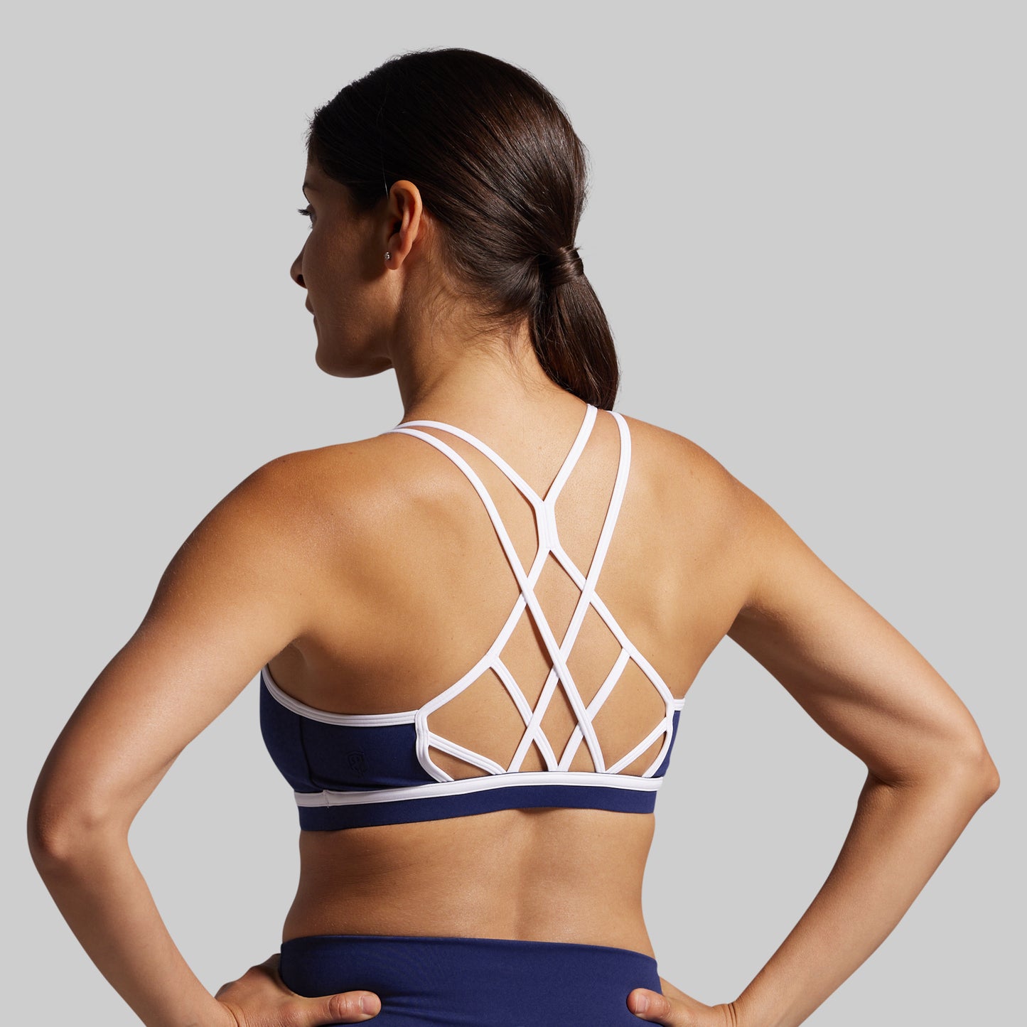 Woman wearing a navy sports bra with criss-cross white straps on a gray background