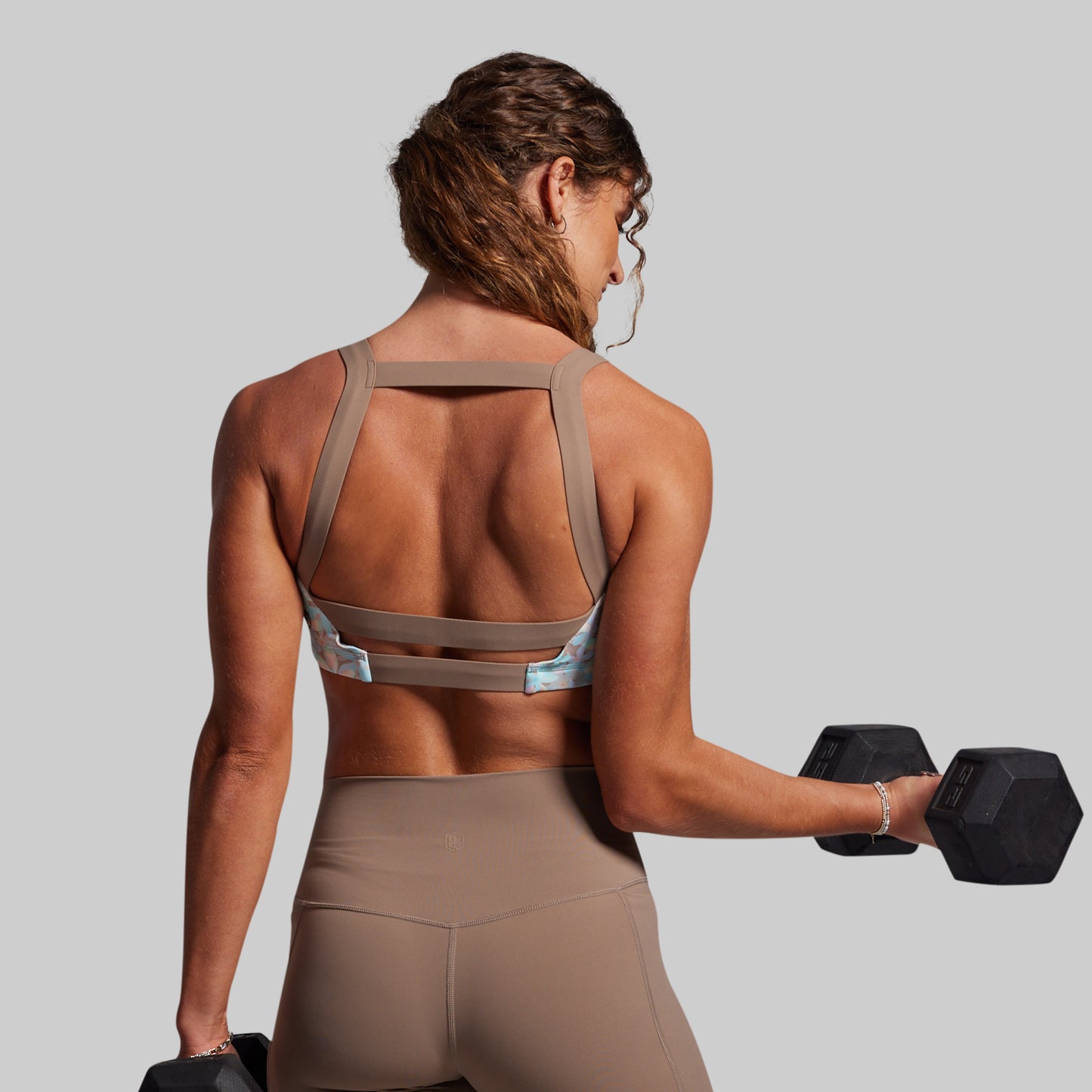 Woman in beige sports bra and leggings holding a black dumbbell against a gray background